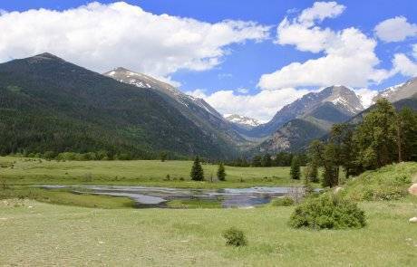 wide shot of calm meadow/tree filled valley with small pond and mountains all around